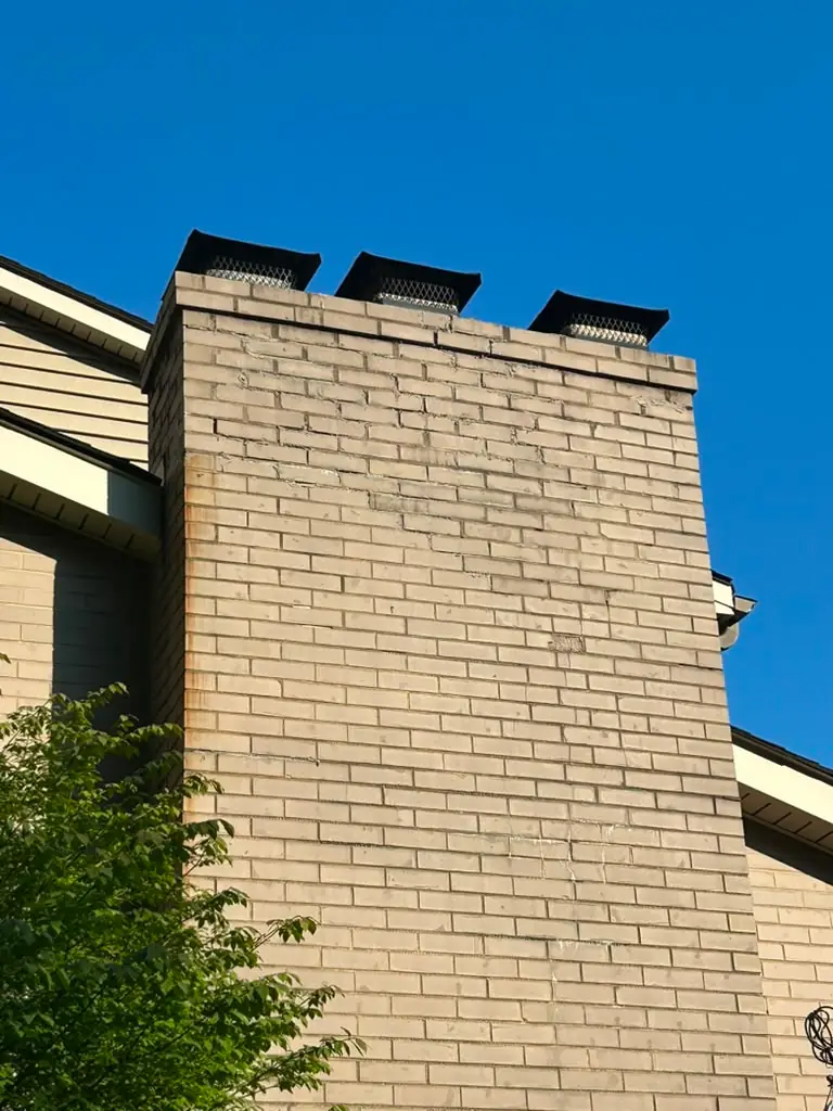 Cracked and open mortar joints stepping through the face of a buff brick chimney in Upper St. Clair, PA, with three capped flues above, before repointing