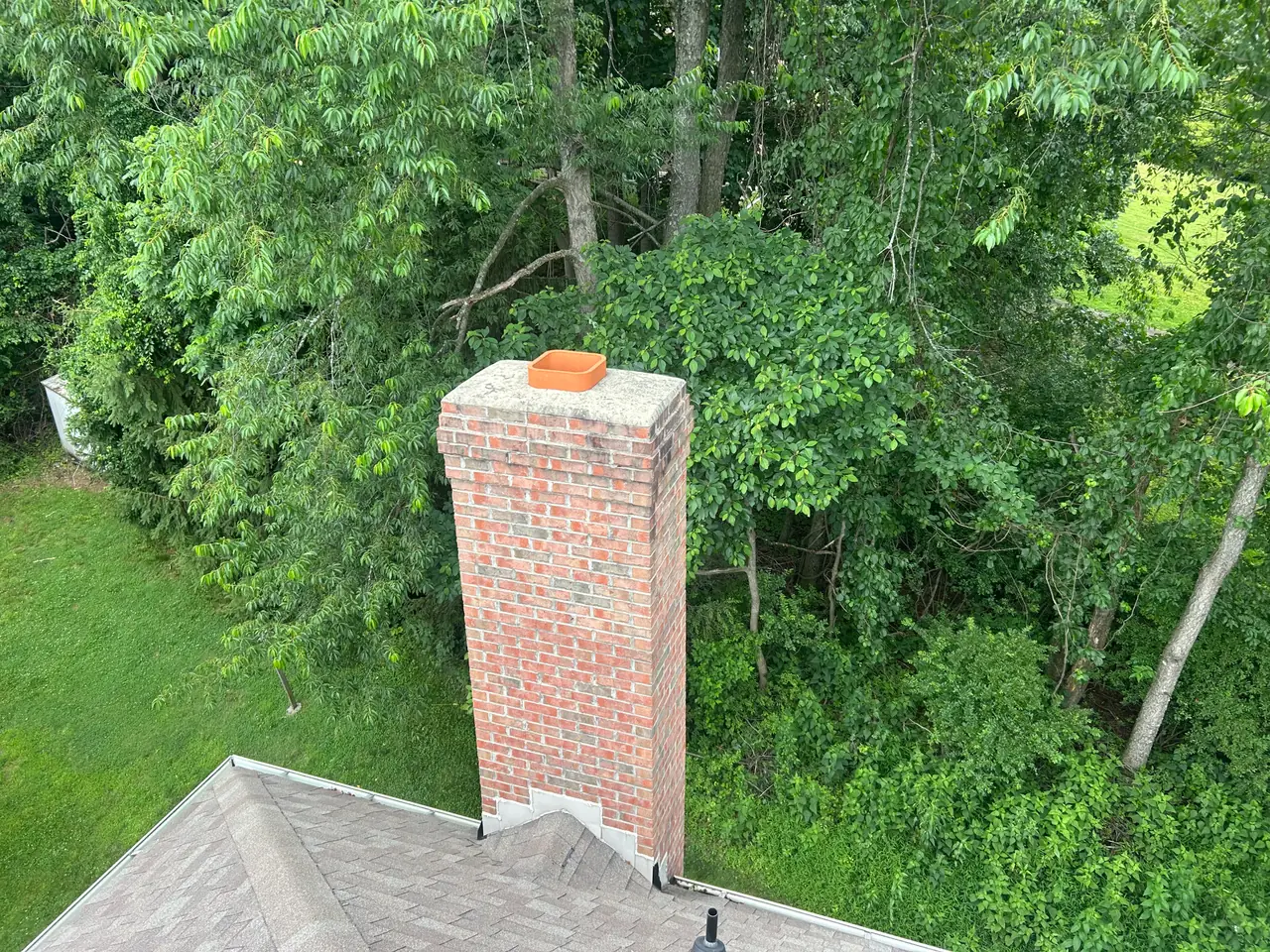 Uncapped clay flue tile and a weathered, cracking crown on a tall brick chimney among the trees in Fox Chapel, PA before repair